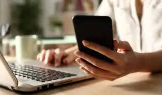 Une main tient un téléphone, l’autre est posée sur le clavier d’un ordinateur portable