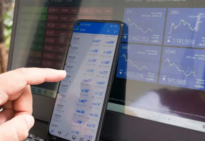 Des cotations de change sur un téléphone, devant l’écran d’une plateforme de trading